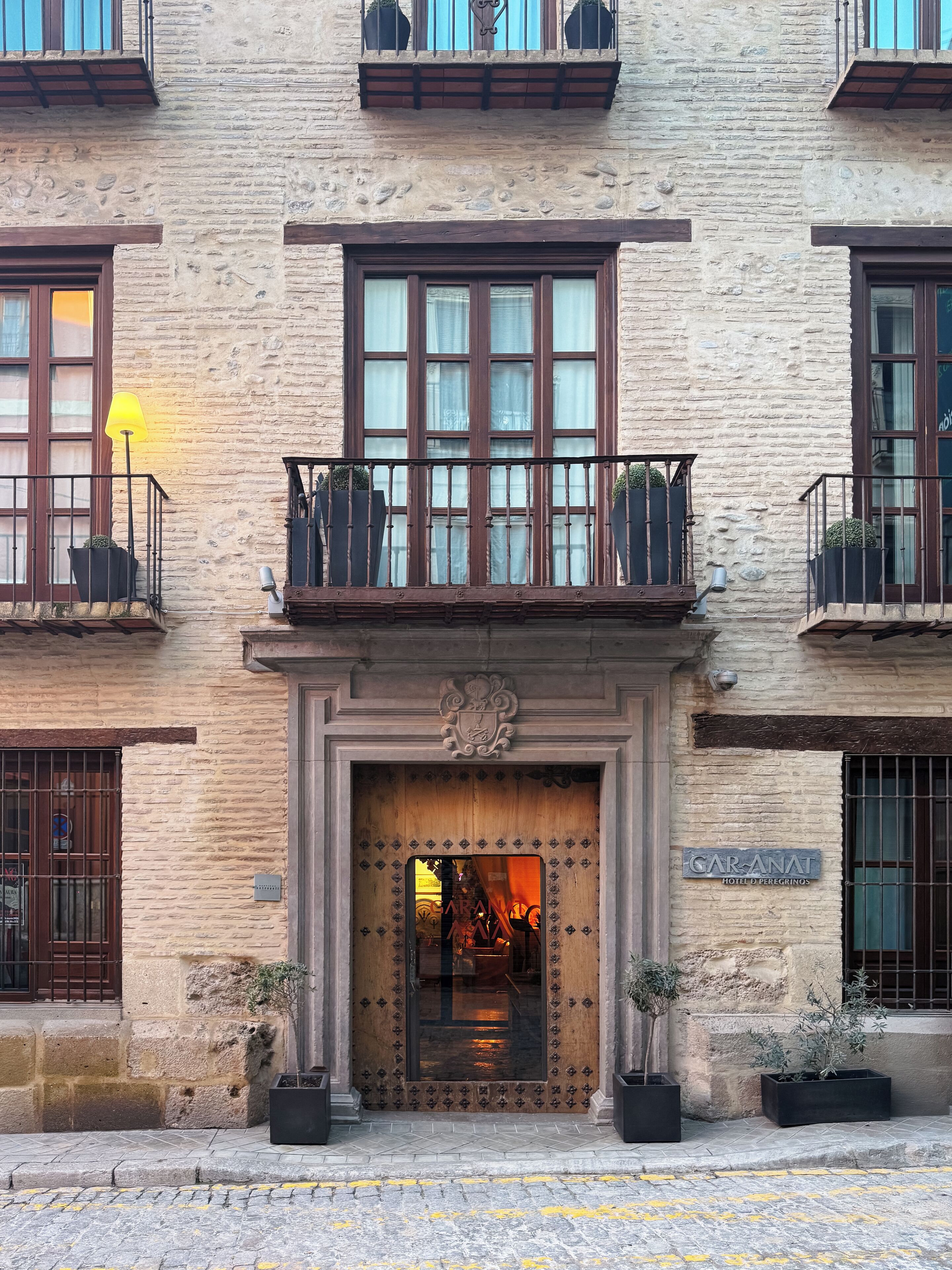 A hotel entrance with a stone facade, a wooden door with a crest, and potted plants on the ground.