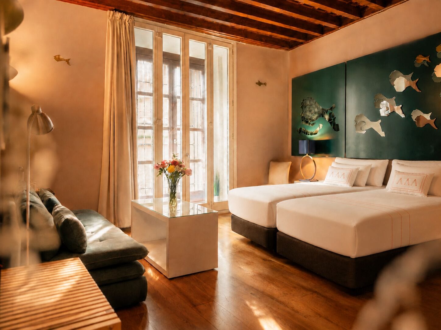A hotel room with a wooden ceiling, a large window with curtains, a sofa, a bed, a small table with a vase of flowers, and a wall with fish decorations.
