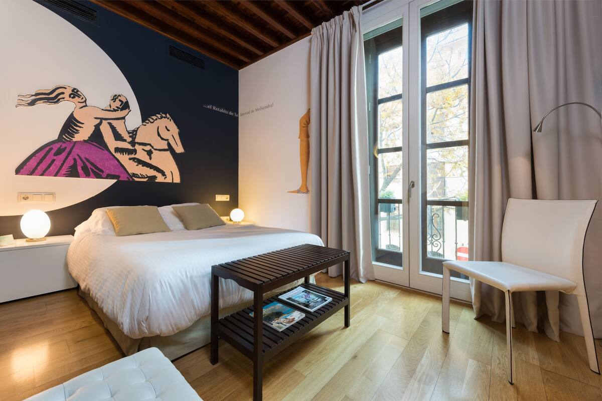 A bedroom with a bed, bedside tables, a reading bench, and a large window with curtains.