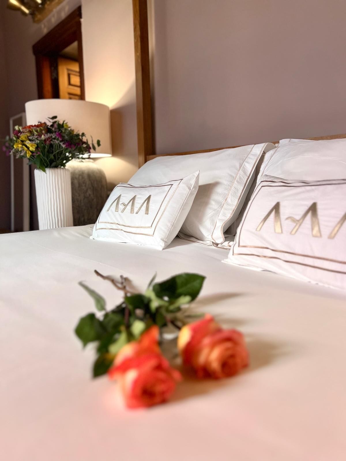 Ein ordentlich gemachtes Bett mit Kissen mit initialen Monogramm, eine Vase mit Blumen und eine Lampe in einem Zimmer mit einer Holztür und einer Wandleuchte.