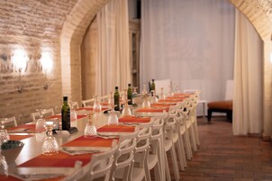 A long dining table with chairs, set with glassware and plates, in a room with brick walls and a tiled floor.