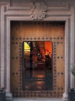 Puerta de madera con un panel de vidrio, un escudo de armas en la parte superior y el reflejo de luces interiores.