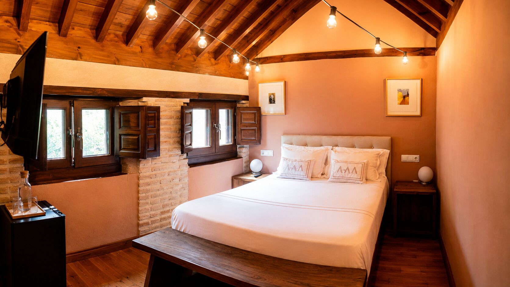 A bedroom with a large bed, wooden headboard, and a brick wall. There are two bedside tables with lamps, a wooden bench, and a TV mounted on the wall.