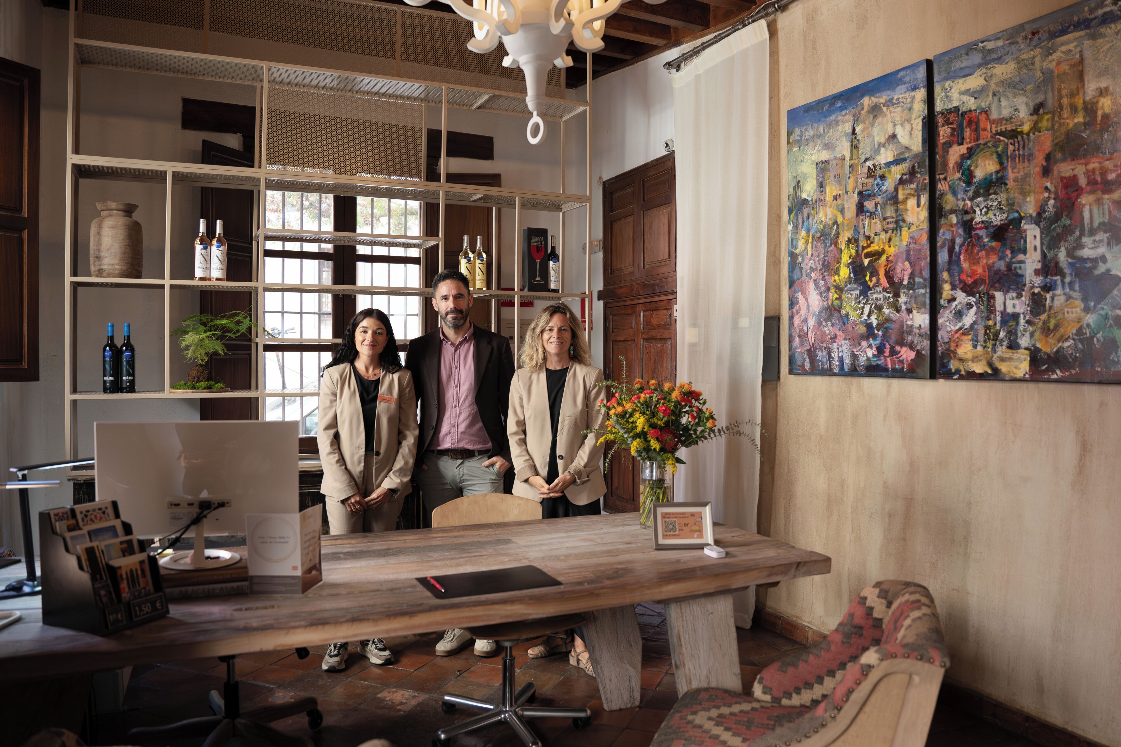 A room with a wooden desk, shelves with bottles, a vase with flowers, and a painting on the wall.