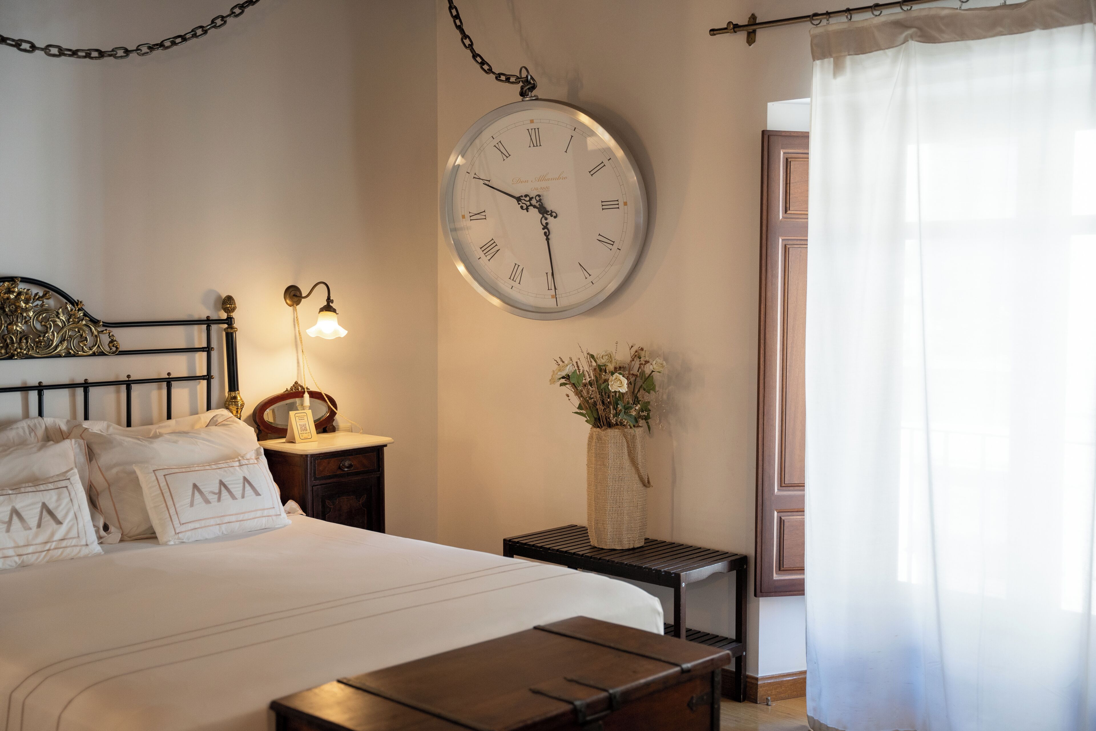 Un dormitorio ordenado con cama, mesita de noche, lámpara, reloj y un florero con flores.