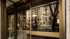 Una ventana de hotel con el nombre "HOTEL PORTA ROMANA" y el reflejo de la calle y edificios a la vista.