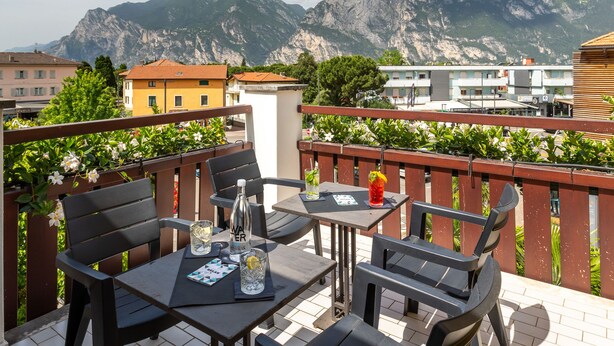 A terrace with a table and chairs, a view of mountains, and buildings in the background. - Hotel Santoni Freelosophy (Nago-Torbole)