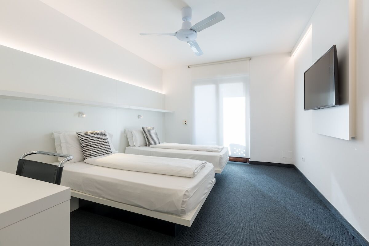 Une chambre d’hôtel moderne équipée de deux lits simples, d’un bureau, d’une chaise, d’un ventilateur de plafond et d’un téléviseur fixé au mur.