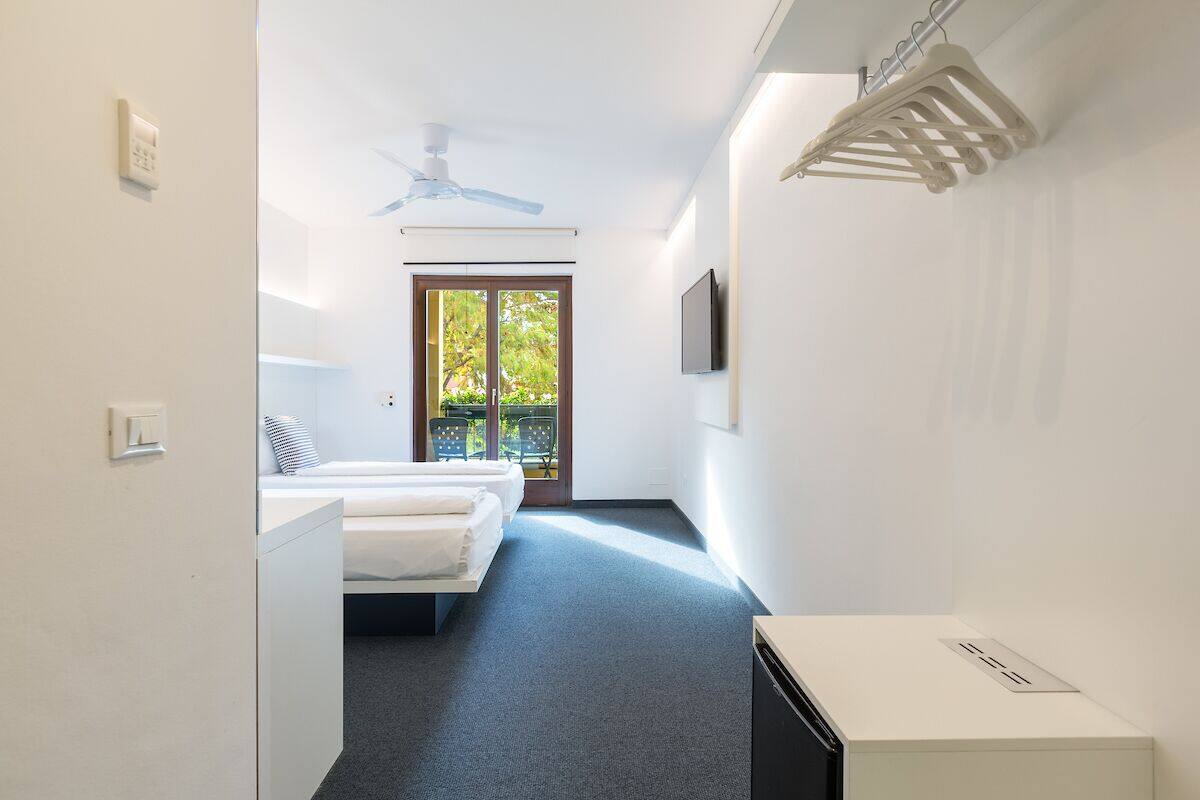 A modern hotel room with a bed, a desk, a ceiling fan, and a view of the outdoors.