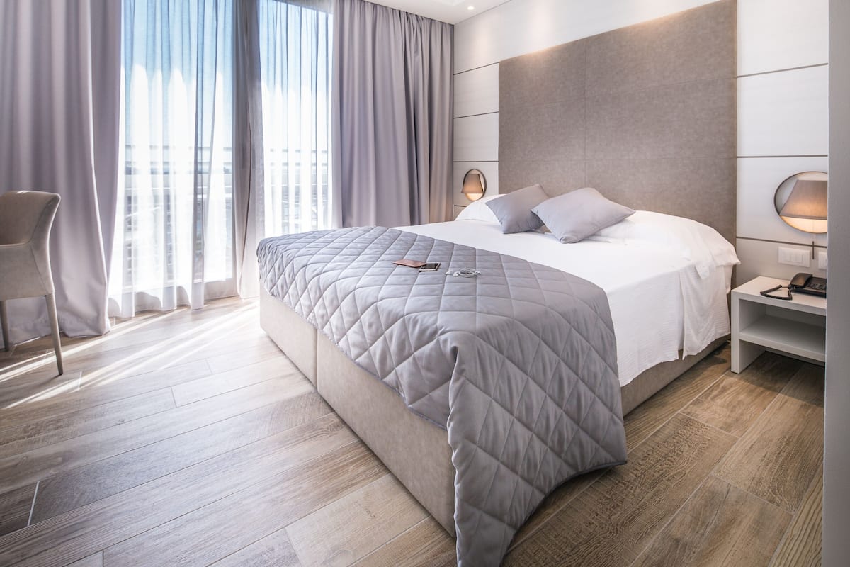 A modern hotel room with a large bed, a bedside table, a chair, and a view of the city through large windows.