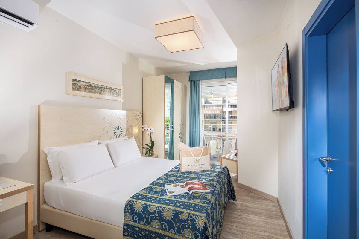 A hotel room with a bed, a bedside table, a nightstand, a window with curtains, a blue door, and a framed picture on the wall.