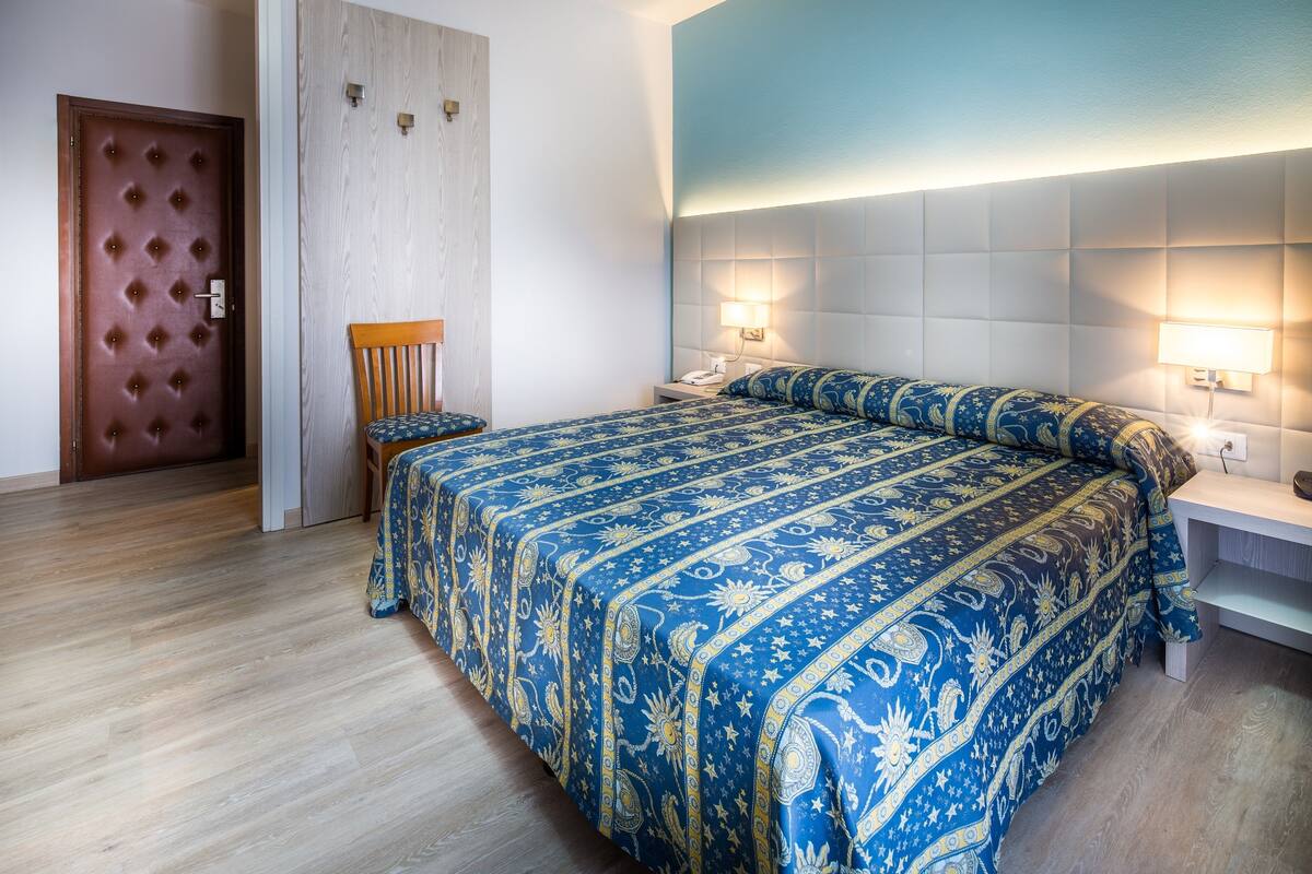 A hotel room with a large bed, a wooden chair, a wooden door, and a nightstand with a lamp.