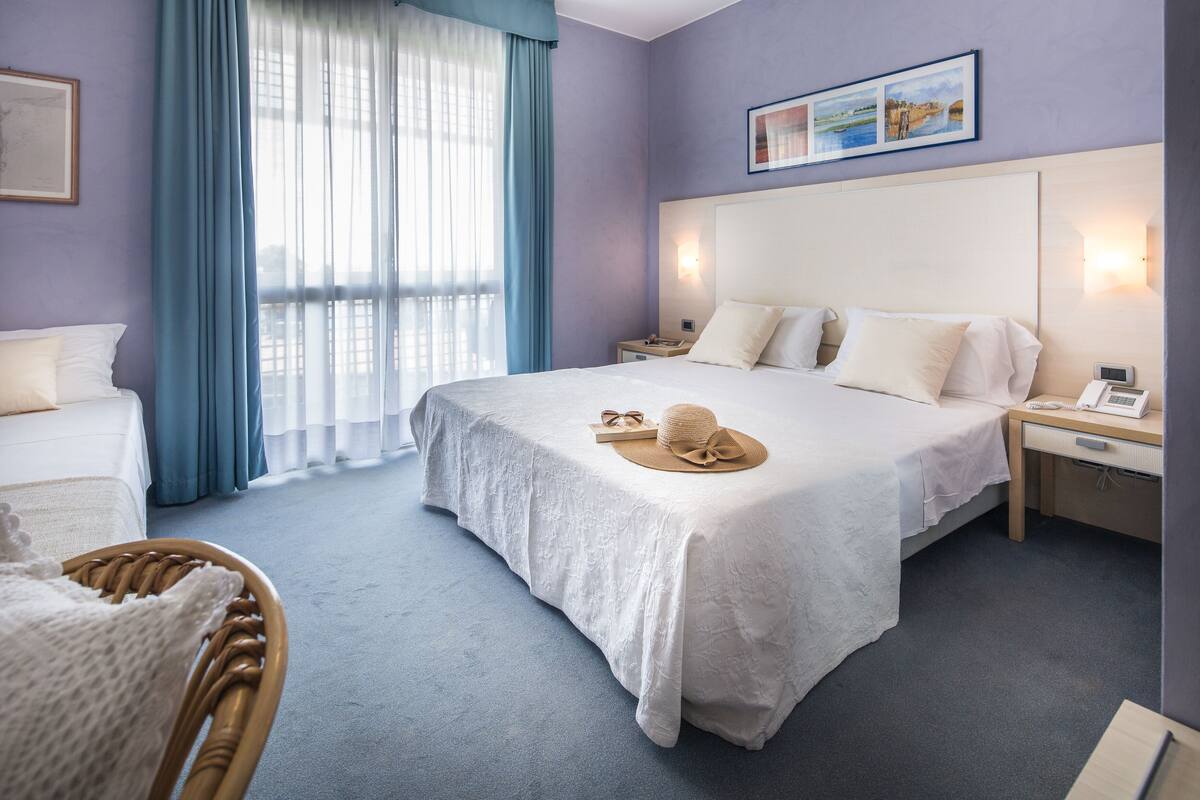 A hotel room with a large bed, a bedside table with a lamp, a chair, and a window with curtains.