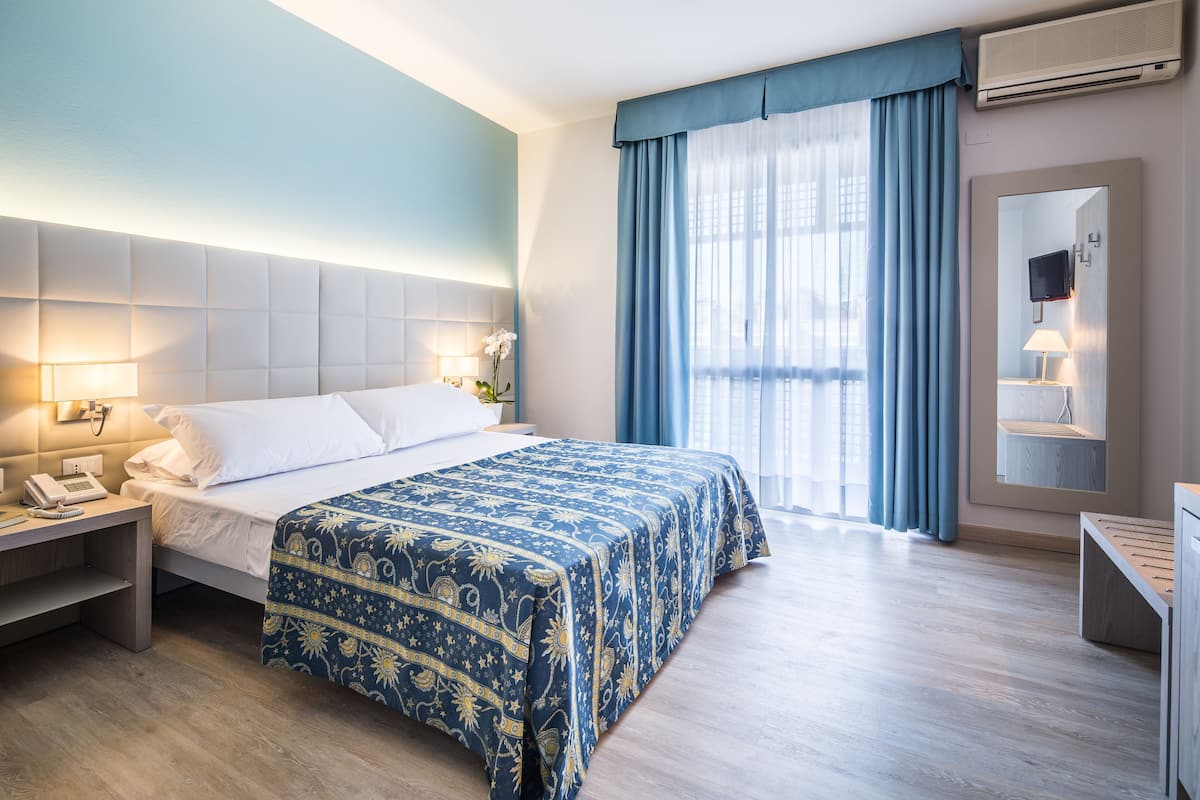 A hotel room with a bed, bedside table, lamp, and a large window with curtains.