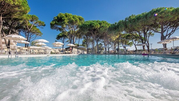 A pool with clear water, surrounded by lounge chairs and umbrellas, and trees in the background. - Hotel San Giorgio (Caorle)