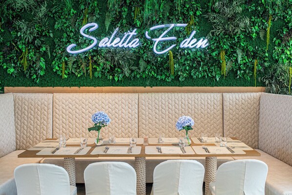 Un restaurante con una pared verde y el nombre 'Seletta Eden' en luces de neón. Hay mesas con sillas blancas y centros de mesa con flores azules.
