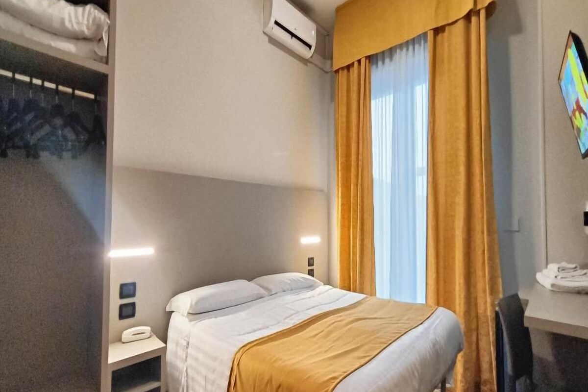 Habitación de hotel con cama, unidad de aire acondicionado, escritorio con silla y ventana con cortinas.