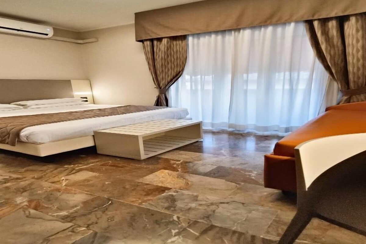 A hotel room with a bed, a bench, a sofa, a large window with curtains, and a ceiling light.