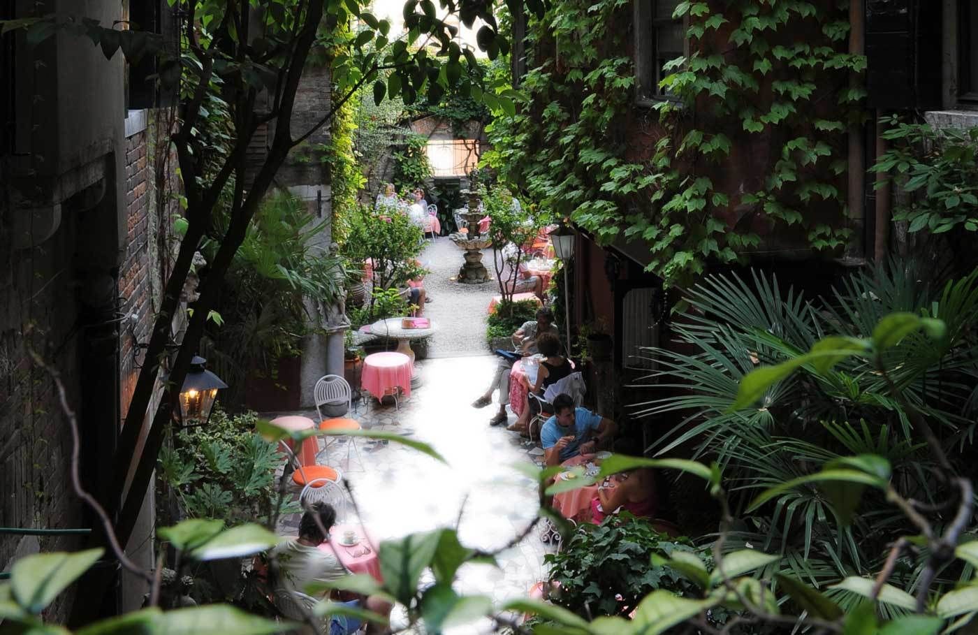 A narrow alley with tables and chairs, surrounded by lush greenery and brick walls.