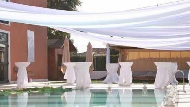 A pool area with white tables and chairs, a clear pool, and a building with a red facade. - Hotel Villa Altura (Ospedaletto Euganeo)