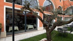 A building with a glass facade, outdoor seating, and a tree in the foreground. - Hotel Villa Altura (Ospedaletto Euganeo)