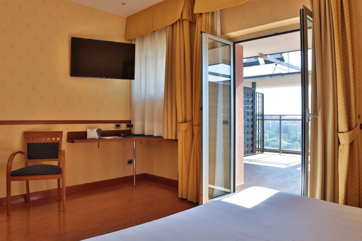 Una camera d'albergo con un letto, una TV a muro, una scrivania con una sedia e un balcone con tende.