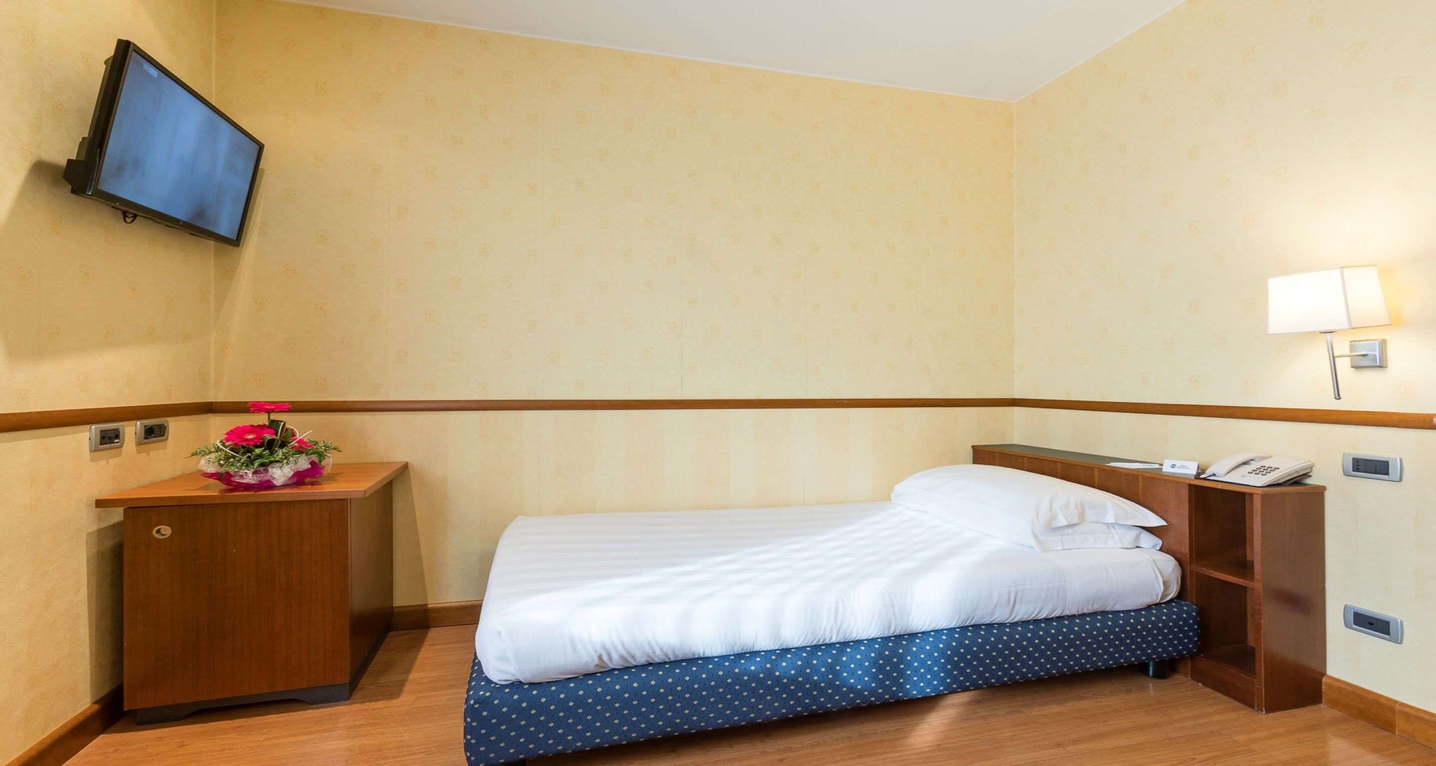 A hotel room with a bed, a wooden nightstand with a telephone, a small desk with a flower vase, and a wall-mounted TV.