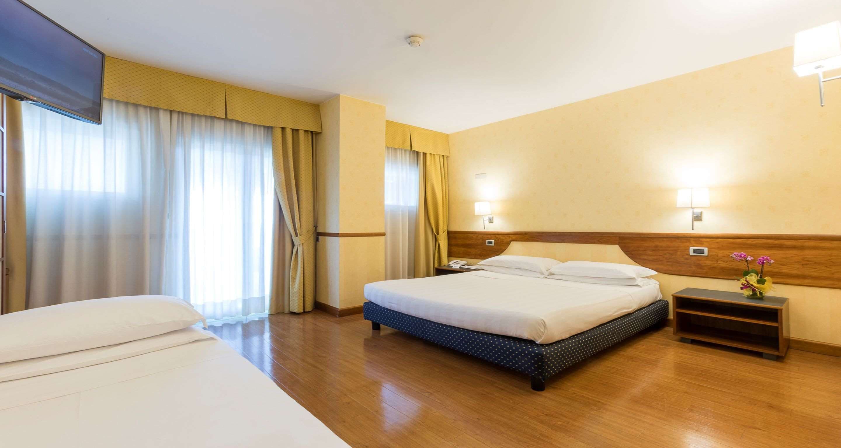 Camera d'albergo con un letto grande, un letto più piccolo, un comodino con una lampada e una testiera in legno.