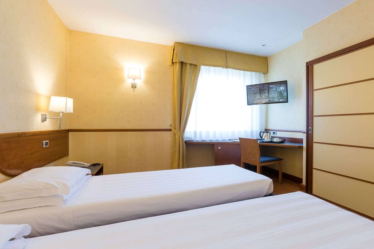 Una camera d'albergo con due letti, una scrivania, una sedia, una lampada e un quadro appeso al muro.