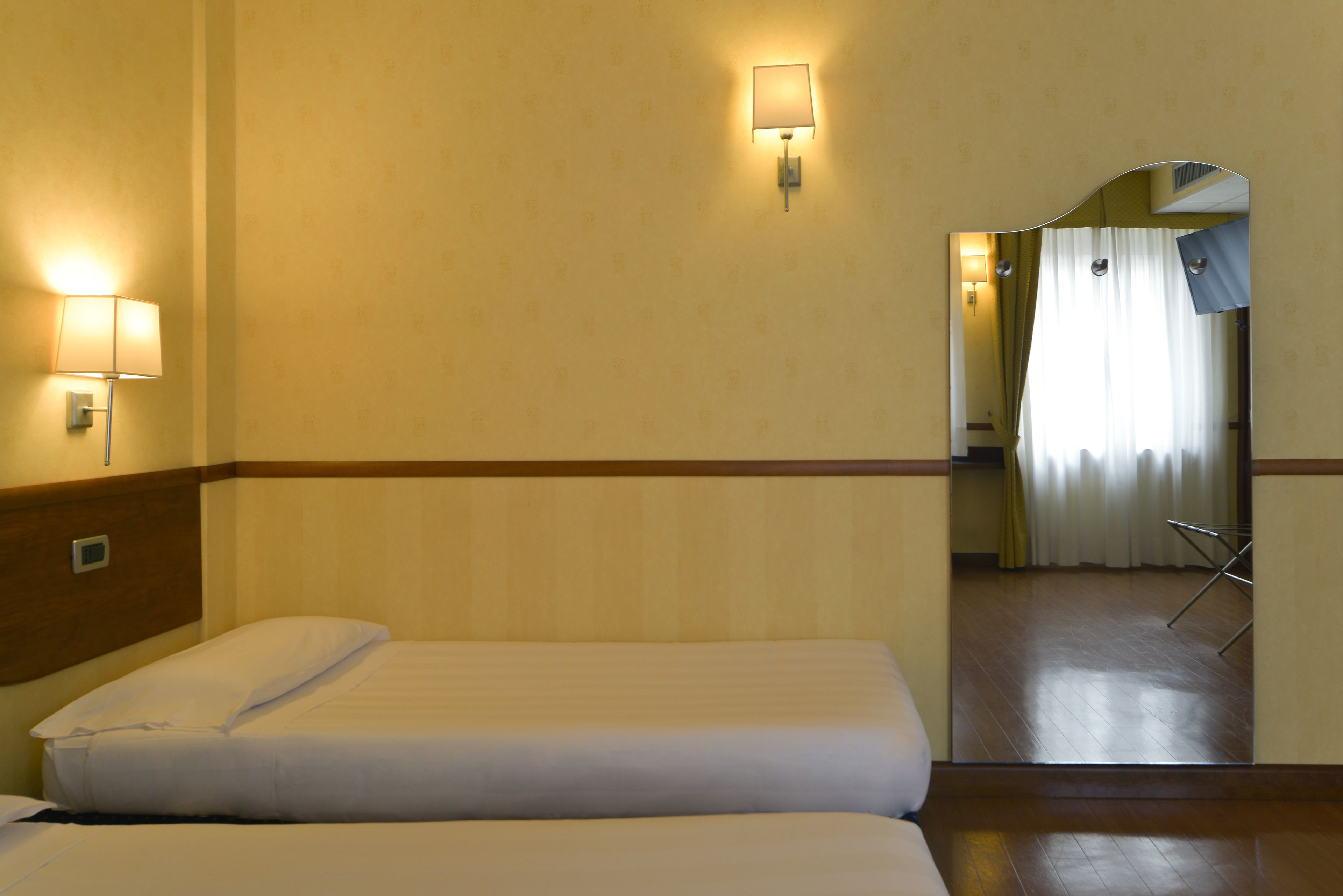 A hotel room with two beds, a nightstand, a wall-mounted lamp, and a mirror reflecting a bathroom with a shower curtain.