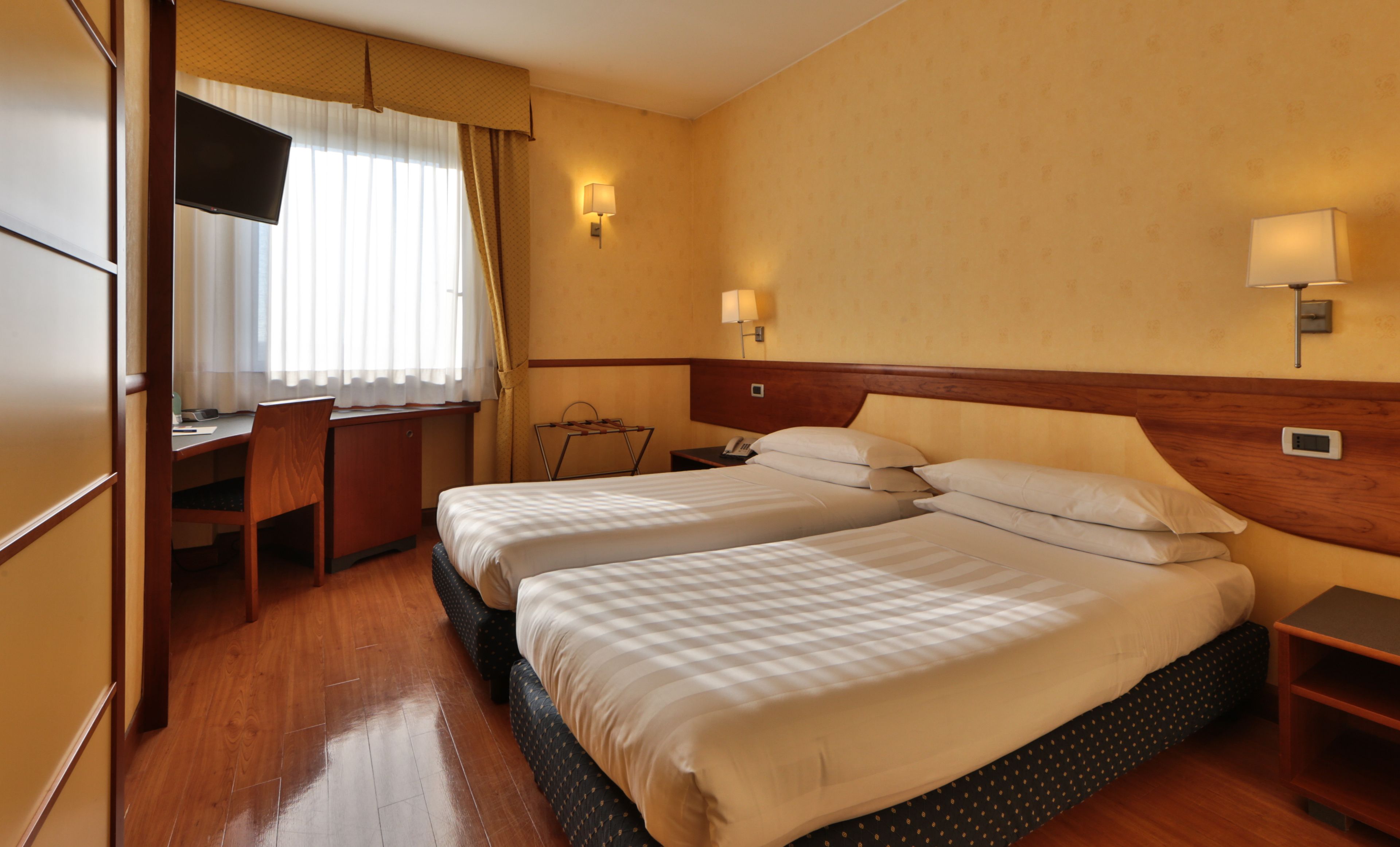 A hotel room with two beds, a desk, a chair, a TV, and two wall lamps.