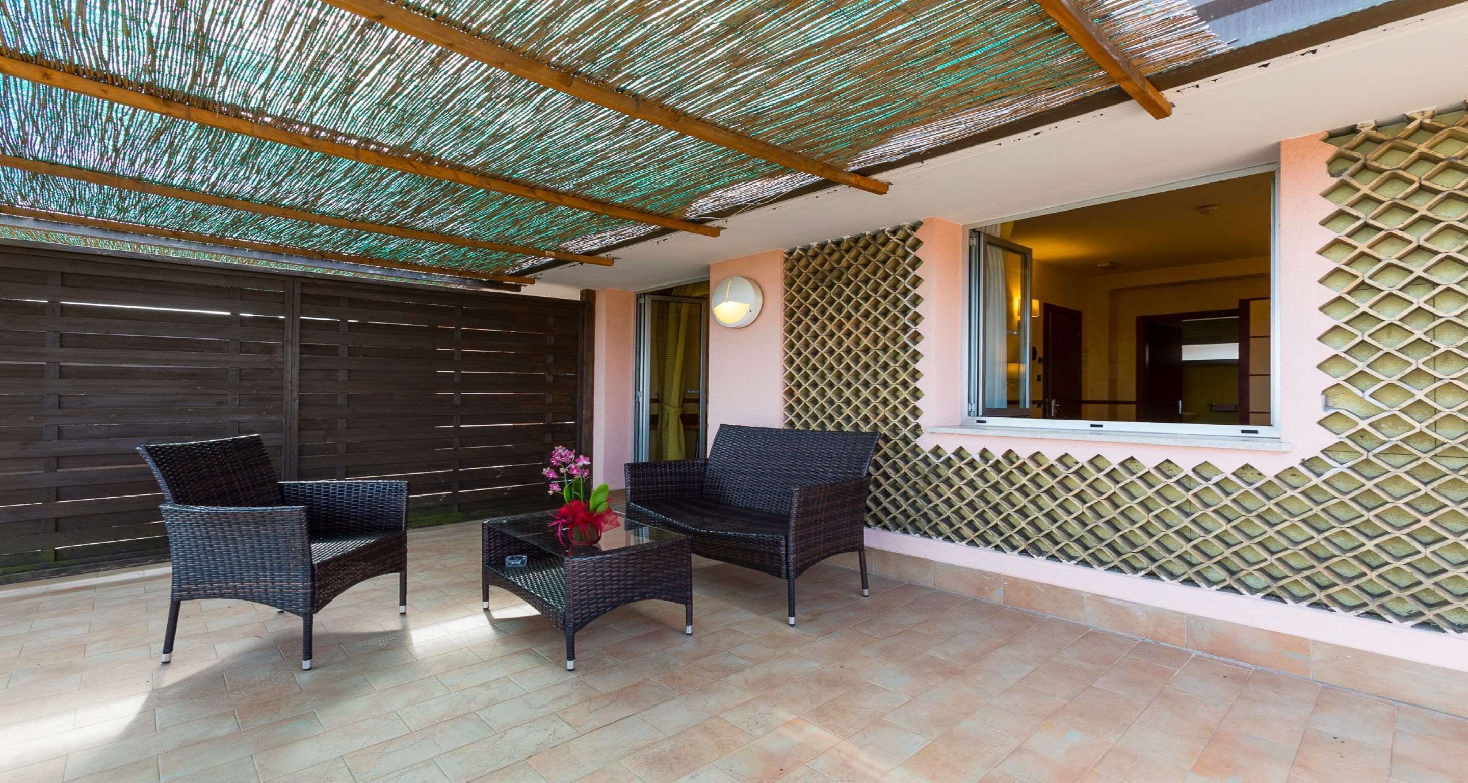 An outdoor patio area with wicker furniture, a glass table with a vase, and a patterned wall.