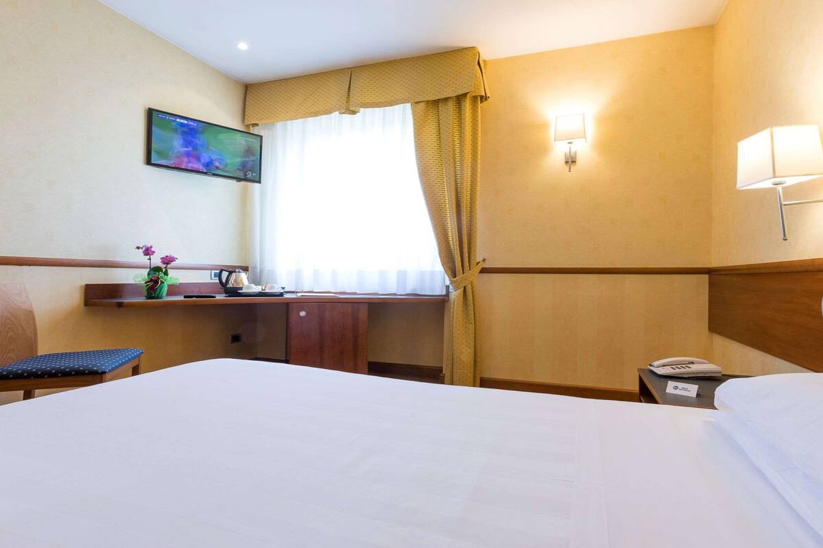 Una camera d'albergo con un letto, una scrivania con una sedia, una TV e una finestra con tende.