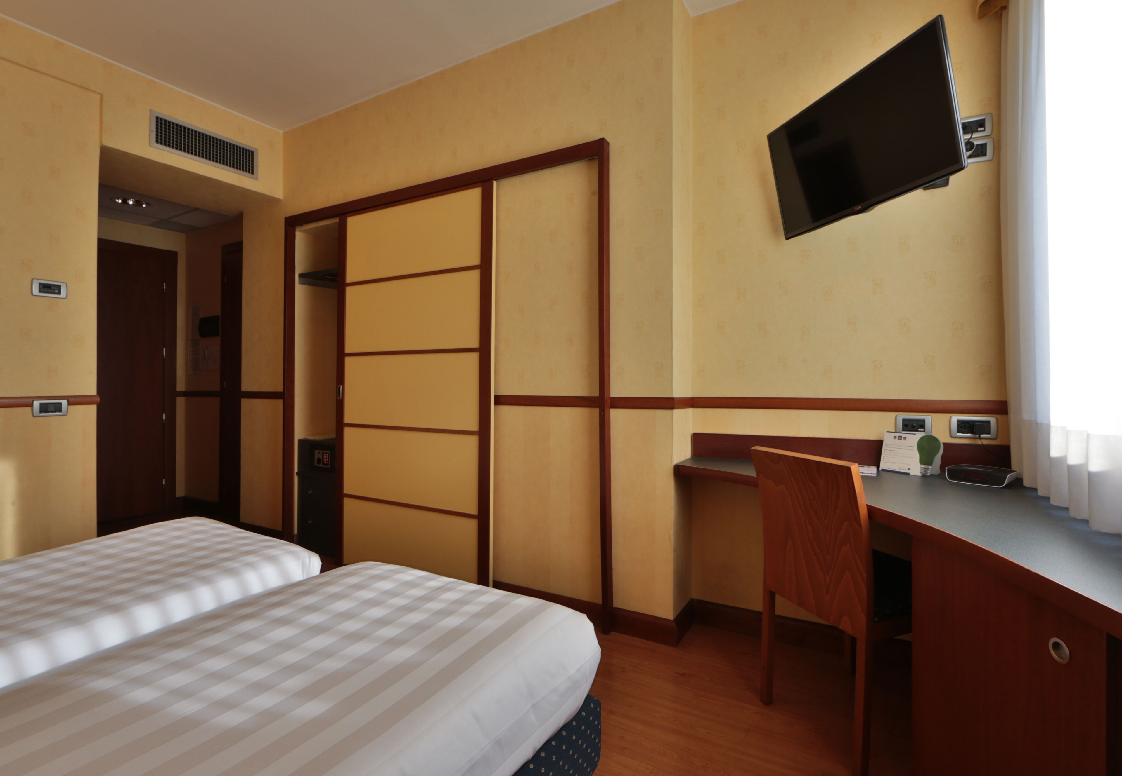 Habitación de hotel con dos camas, un escritorio, una silla, un televisor montado en la pared y una ventana con cortinas.