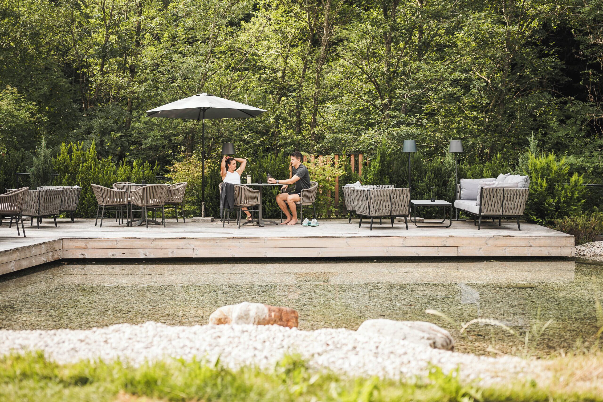 A deck with outdoor furniture, a table, and chairs, surrounded by a natural landscape and a body of water.