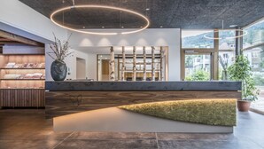 A modern reception area with a wooden reception desk, a large vase with branches, and a view of the surrounding buildings through large windows. - Sporthotel Zoll (Vipiteno)