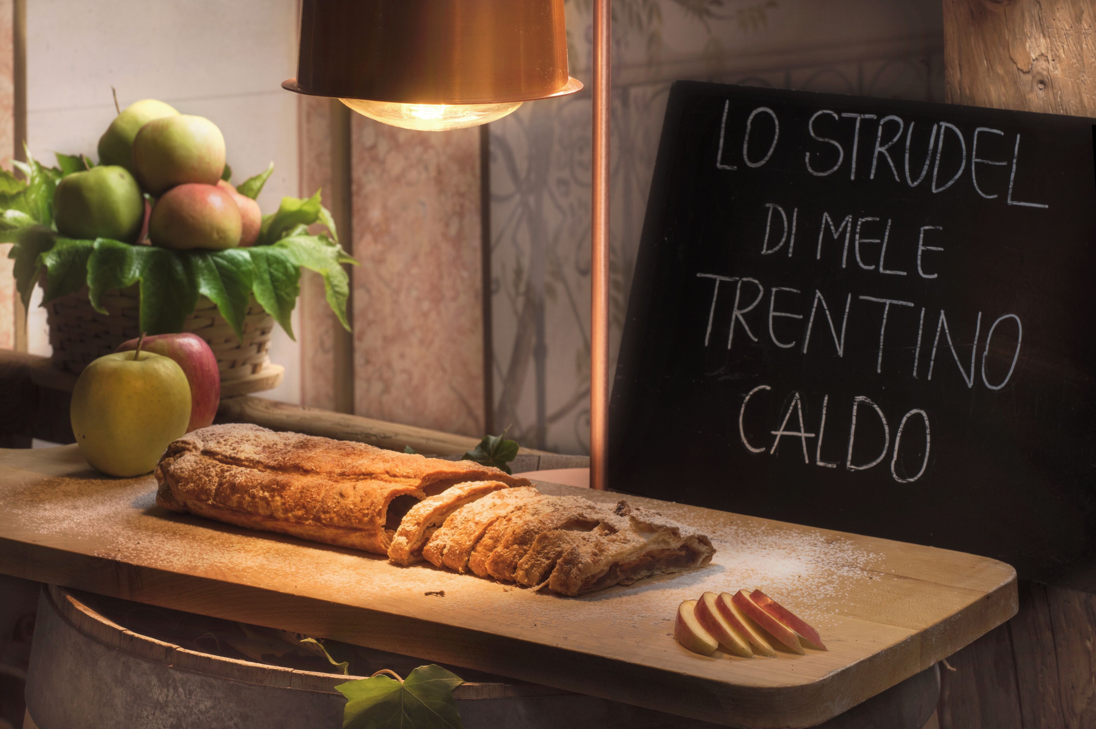 A loaf of bread on a wooden board, a basket of apples, and a sign with Italian text.