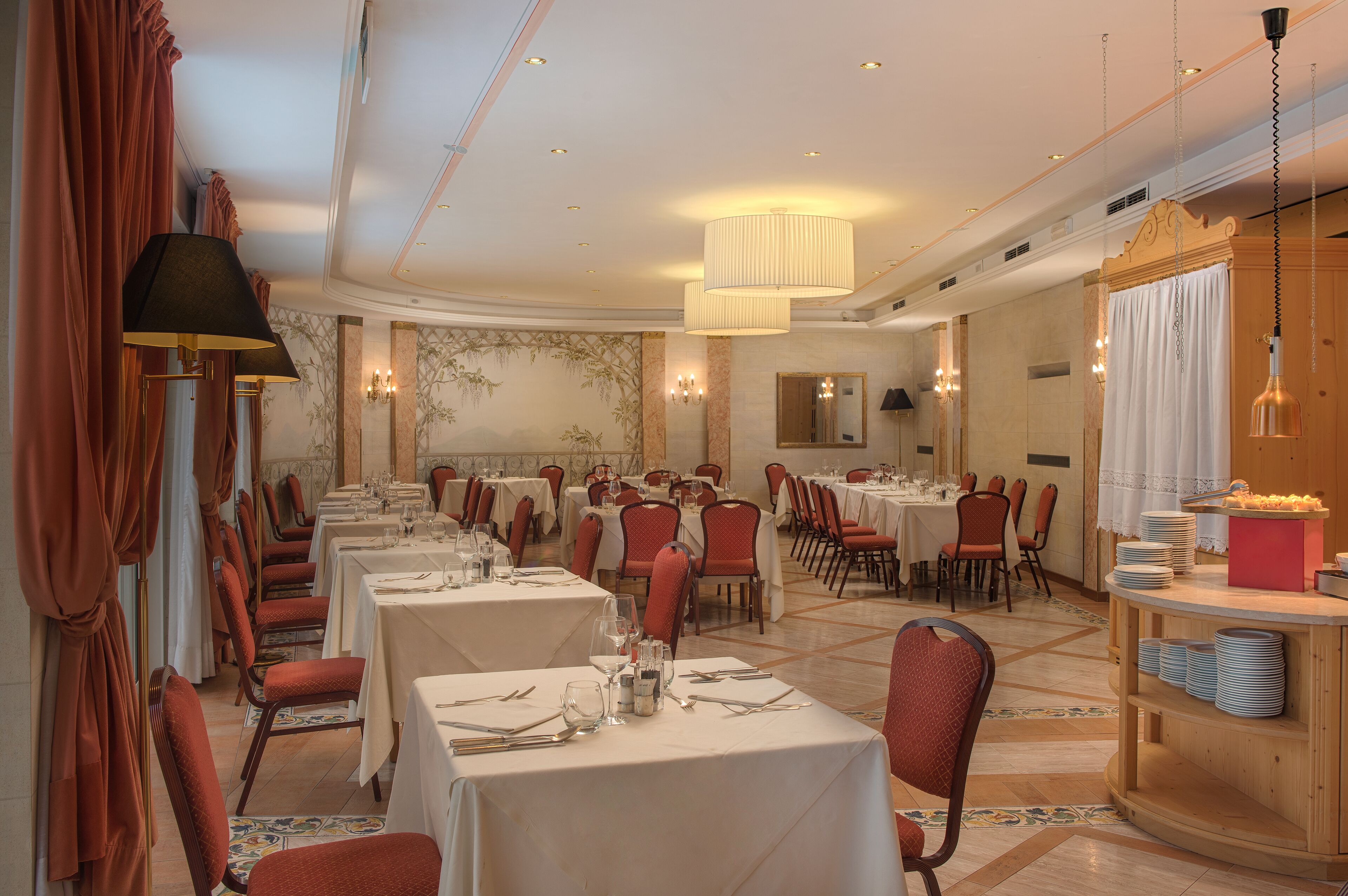 A formal dining room with multiple tables set for service, red upholstered chairs, and a buffet area with stacked plates.