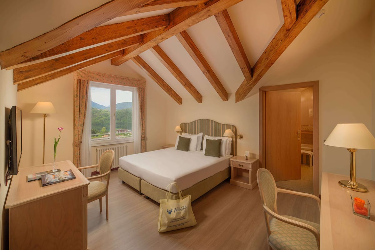 Ein Schlafzimmer mit Holzbalkendecke, einem Bett mit Kissen, einem Schreibtisch mit Lampe und einem Fenster mit Blick ins Grüne.