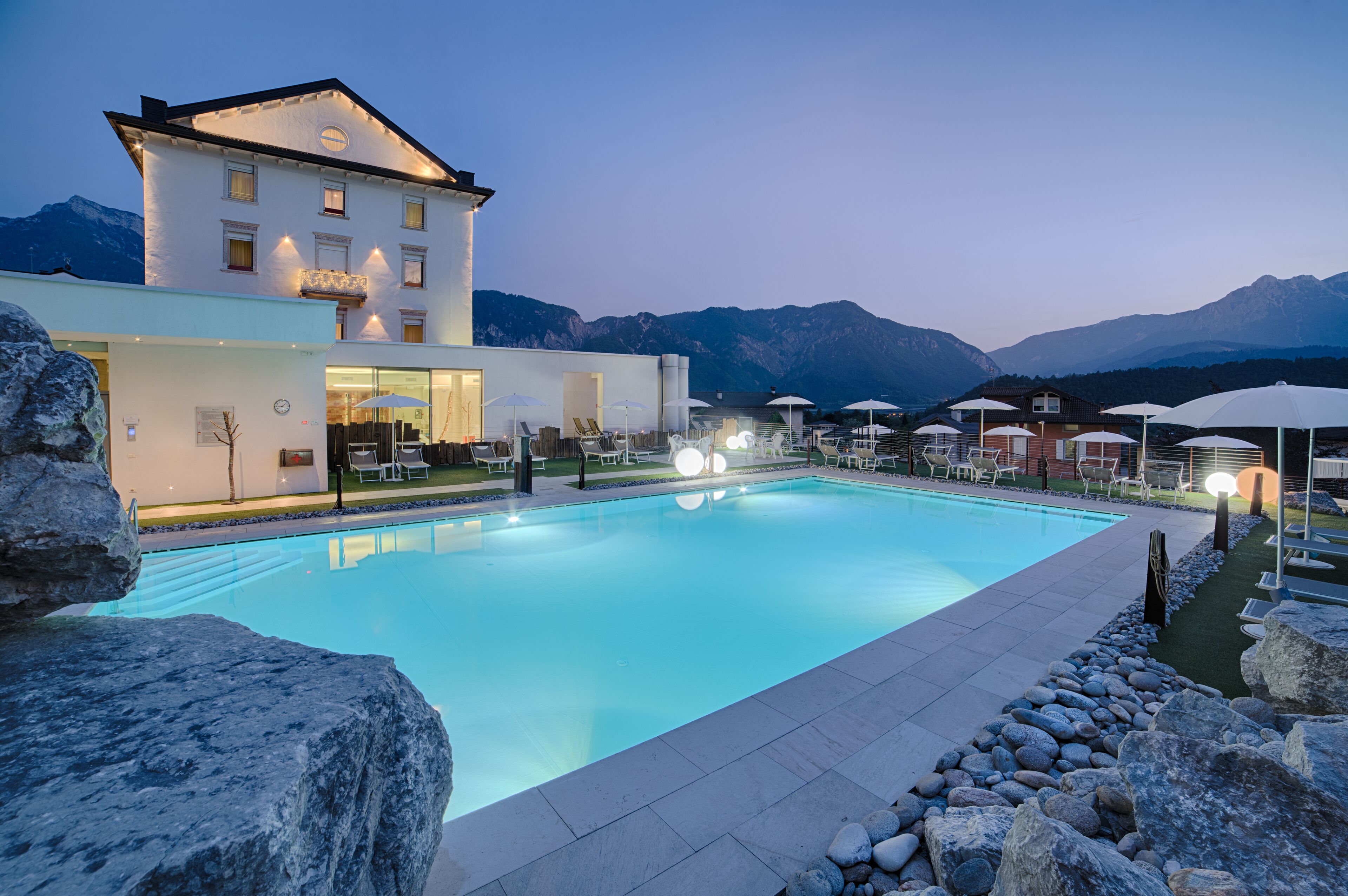 Une piscine avec des chaises longues et des parasols, un bâtiment au design moderne, et des montagnes en arrière-plan.