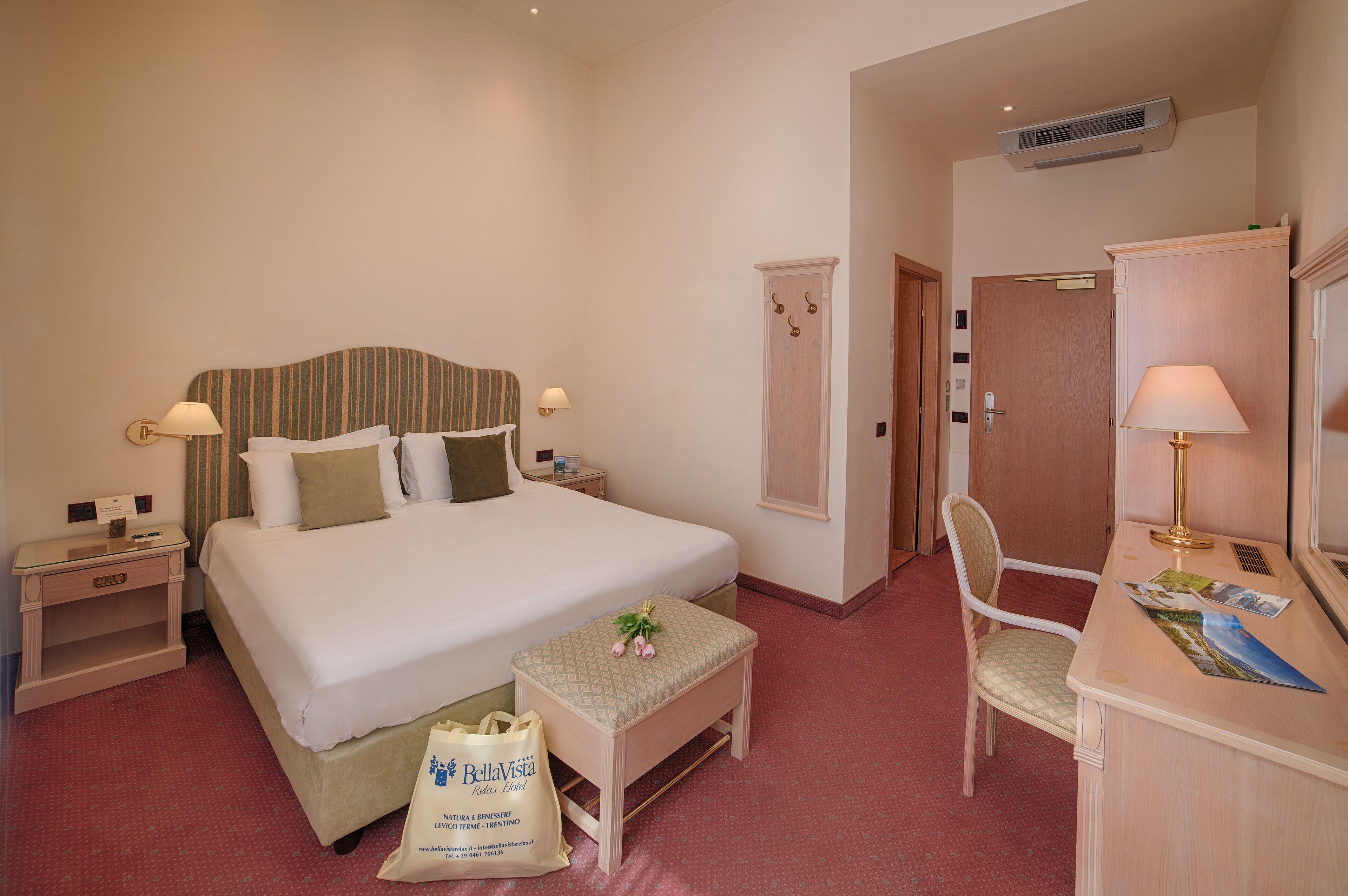 A hotel room with a large bed, bedside tables, a desk with a lamp, a chair, and a wardrobe.