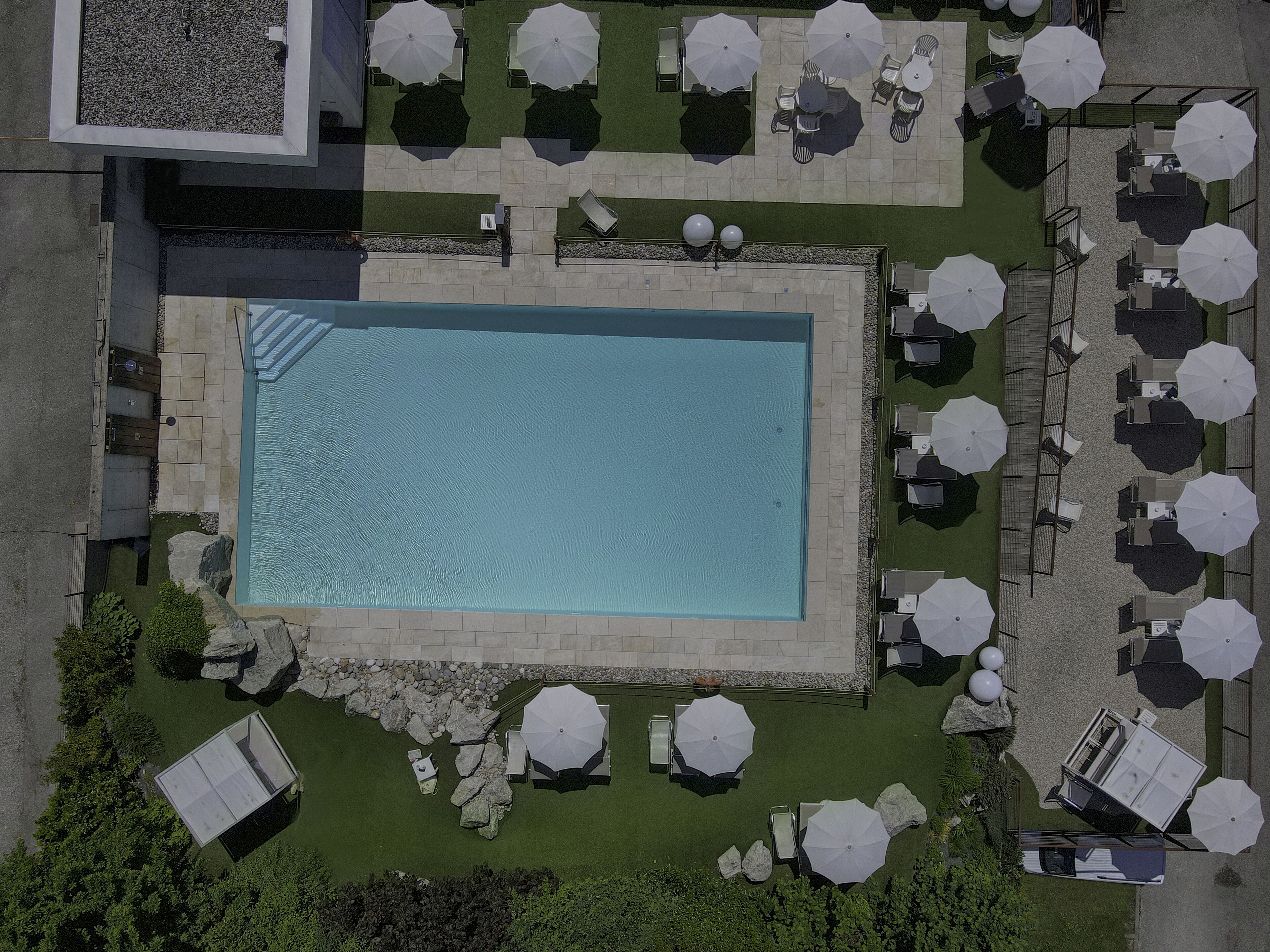 Vista aérea de una piscina rectangular rodeada de sillas de descanso y sombrillas, con un muro de piedra y follaje.