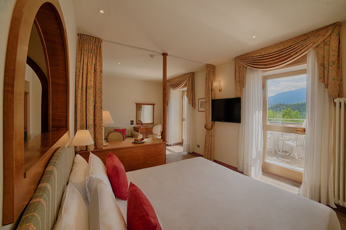 Ein Hotelzimmer mit einem großen Bett, einem hölzernen Kopfteil, einem Schminktisch, einem Spiegel, einem Fernseher und einem Balkon mit Bergblick.