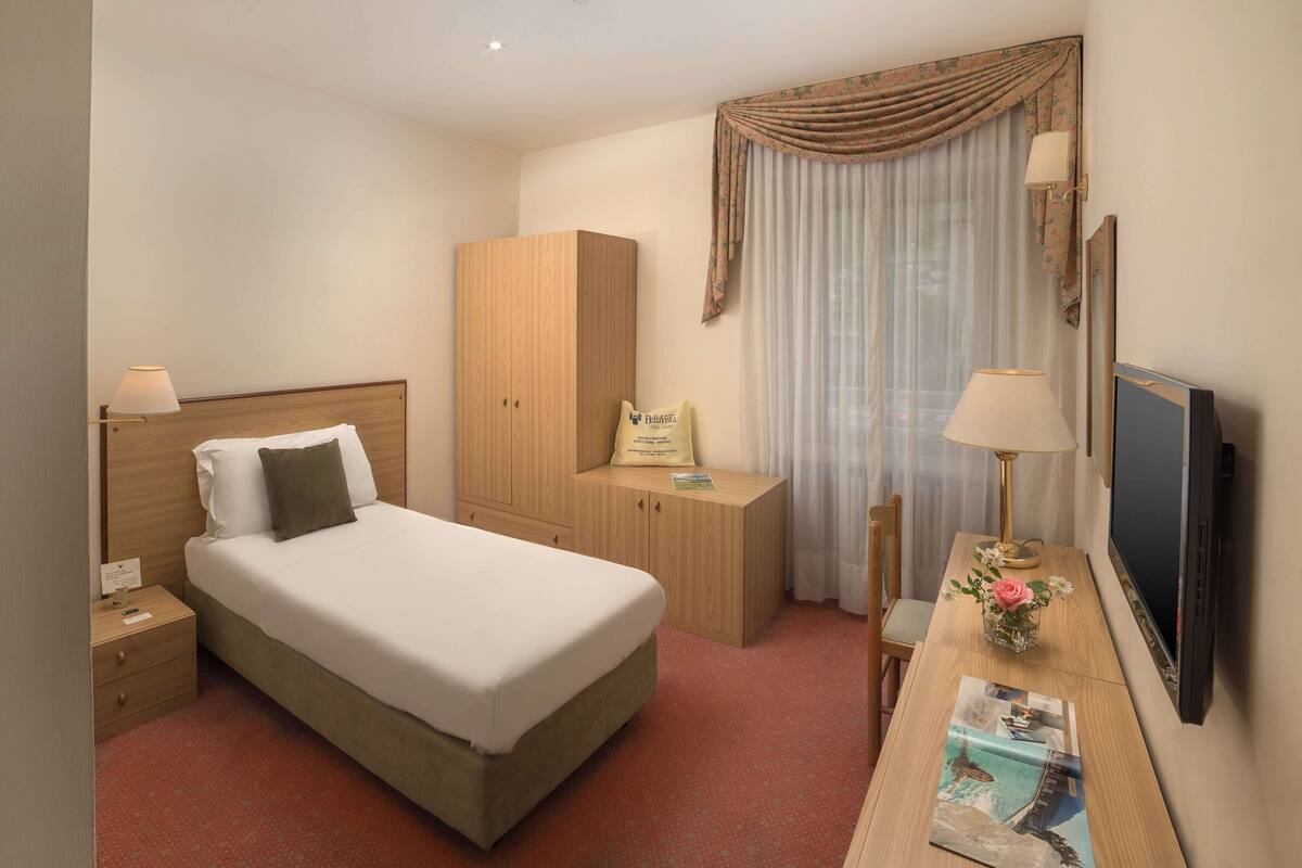 Ein Hotelzimmer mit einem Bett, einem Holzschrank, einem Schreibtisch mit Lampe, einem Fernseher und einem Fenster mit Vorhängen.