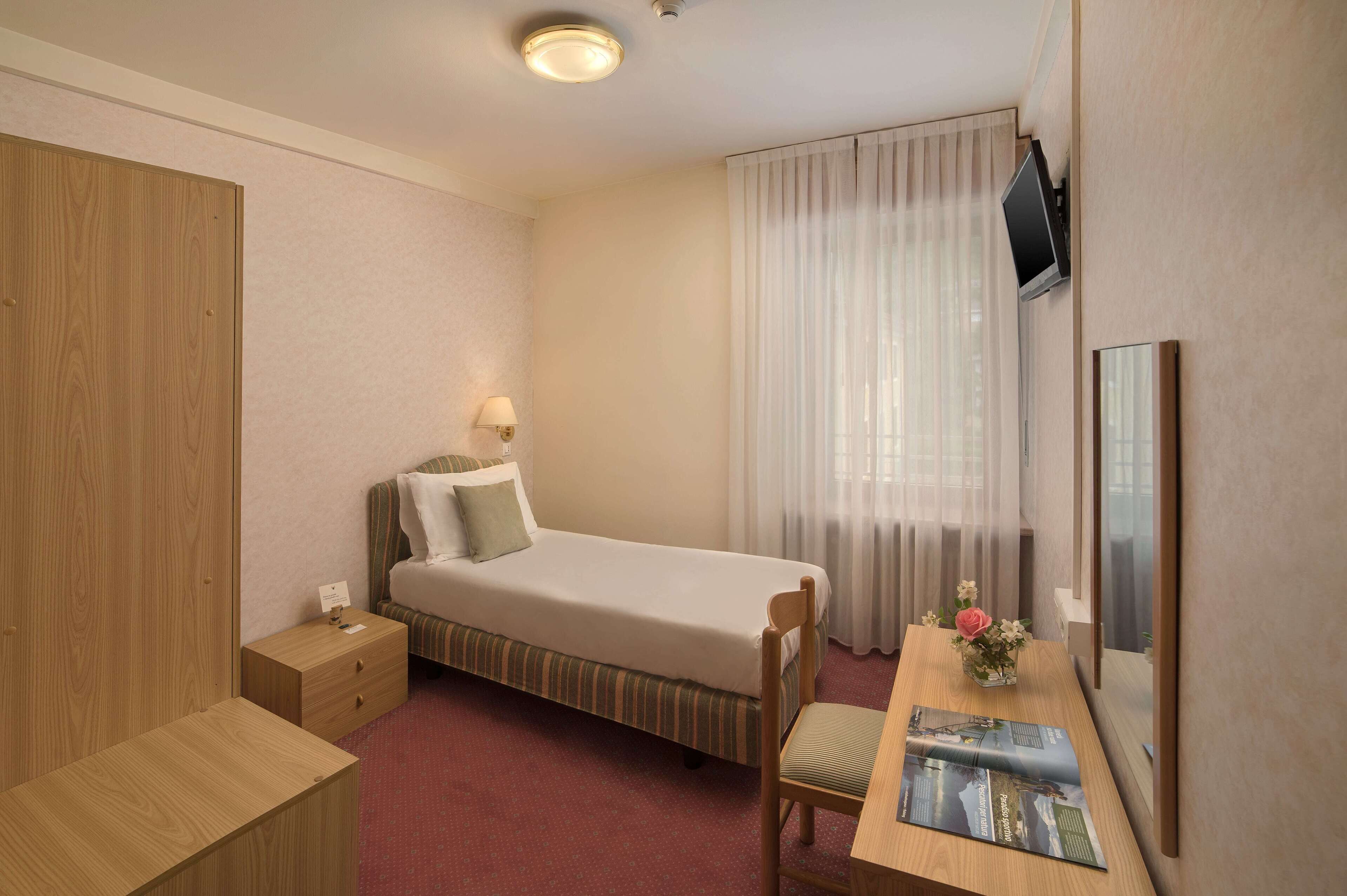 A hotel room with a single bed, a desk with a chair, a nightstand, a TV mounted on the wall, and a window with sheer curtains.