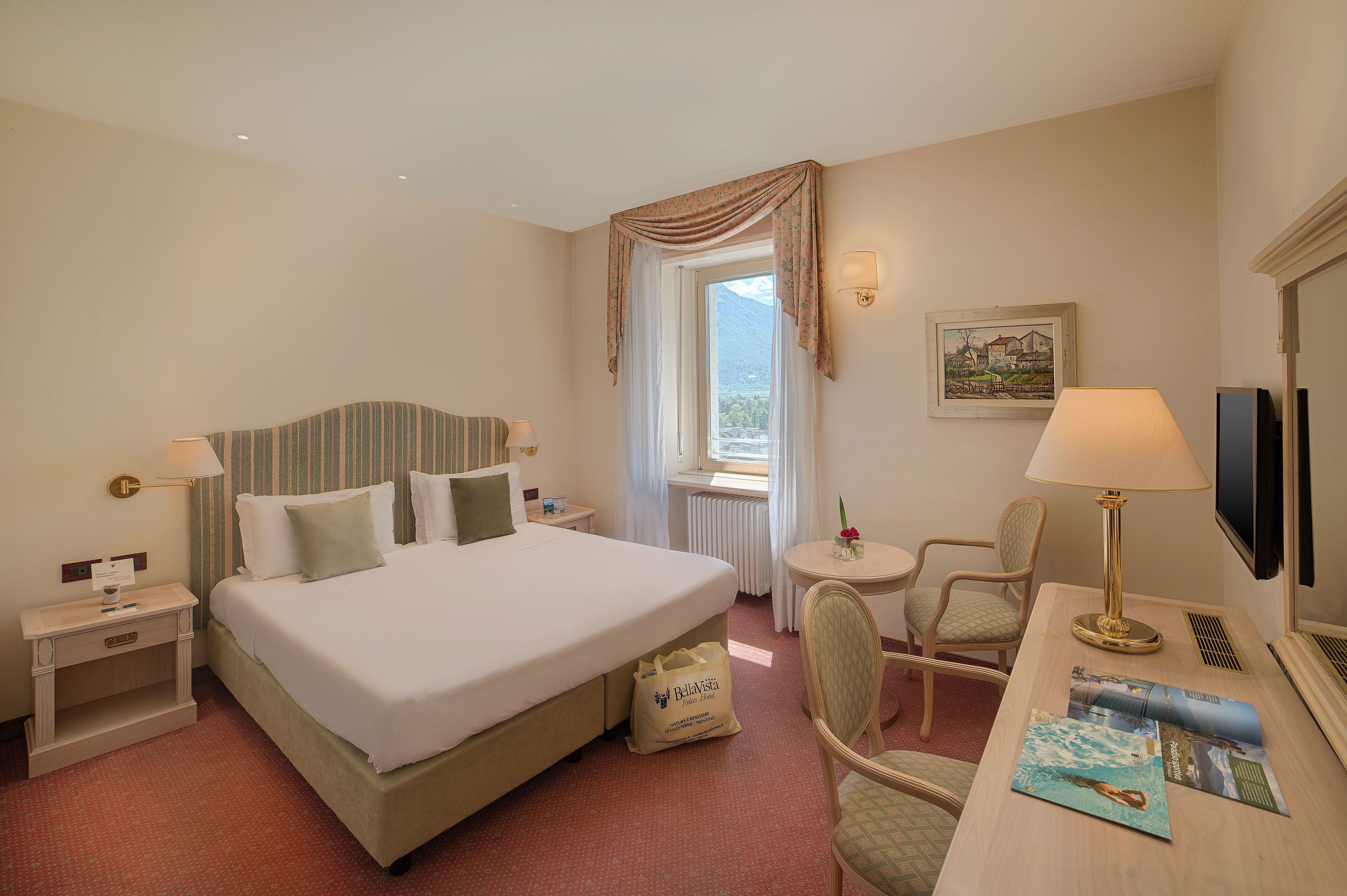 A hotel room with a large bed, a desk with a lamp, a chair, a small table, and a window with curtains.