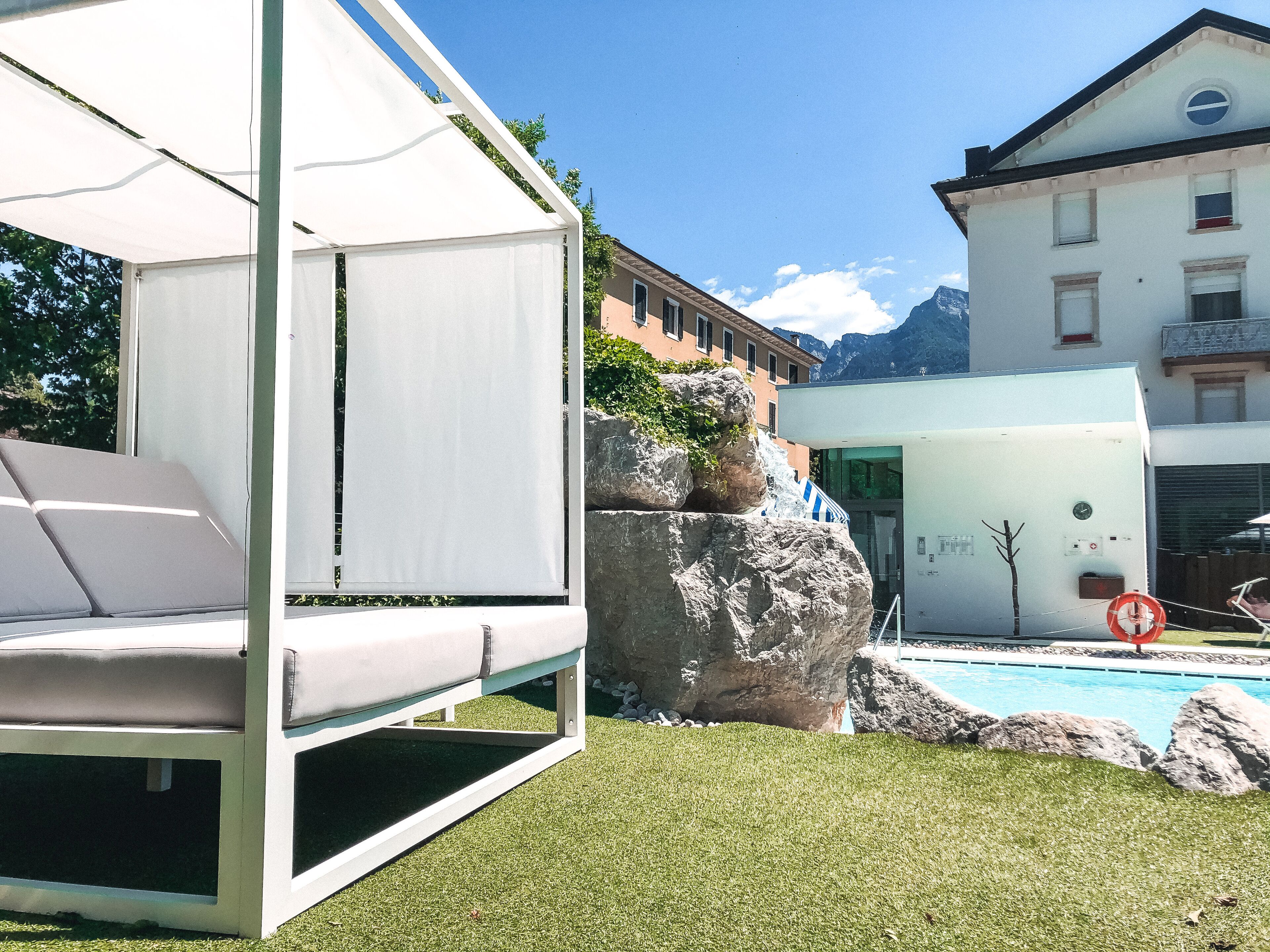 Un espacio moderno para sentarse al aire libre con un toldo blanco, una piscina y un edificio con el paisaje de montañas al fondo.