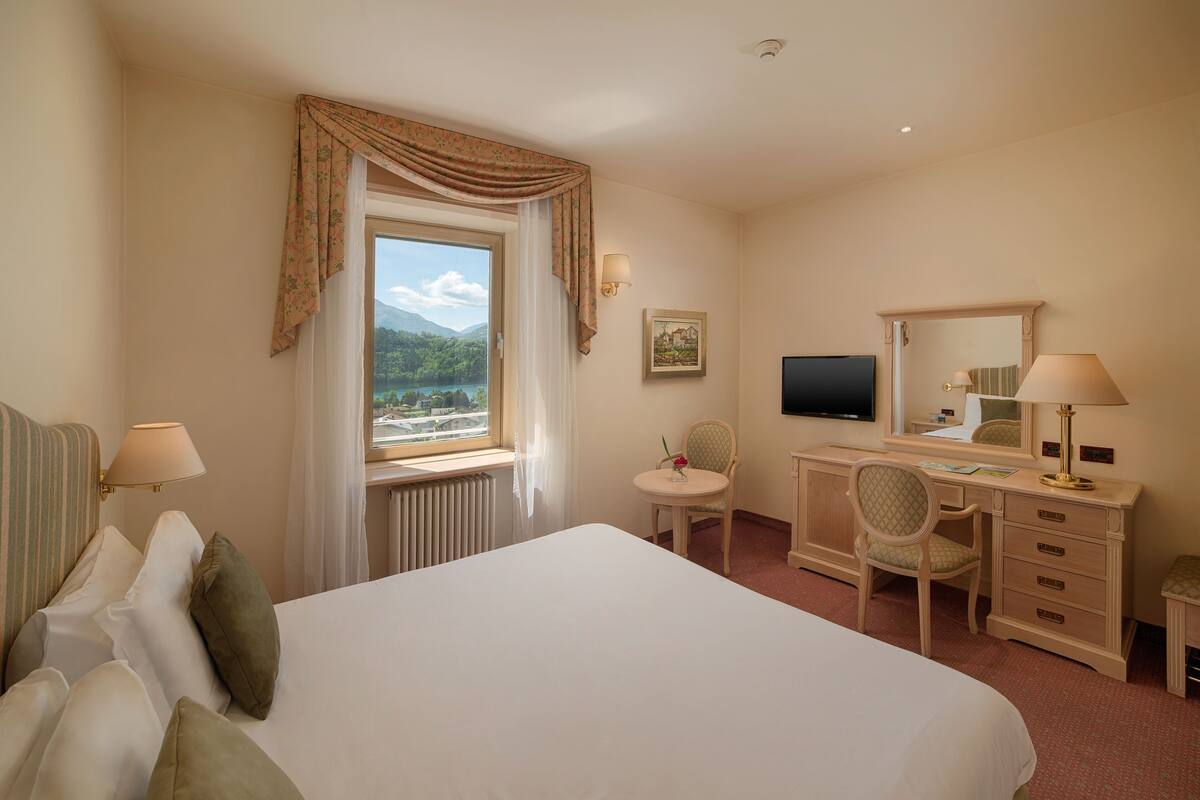 Ein Hotelzimmer mit einem großen Bett, einem Schreibtisch mit Stuhl, einem Fernseher und einem Fenster mit Vorhängen.