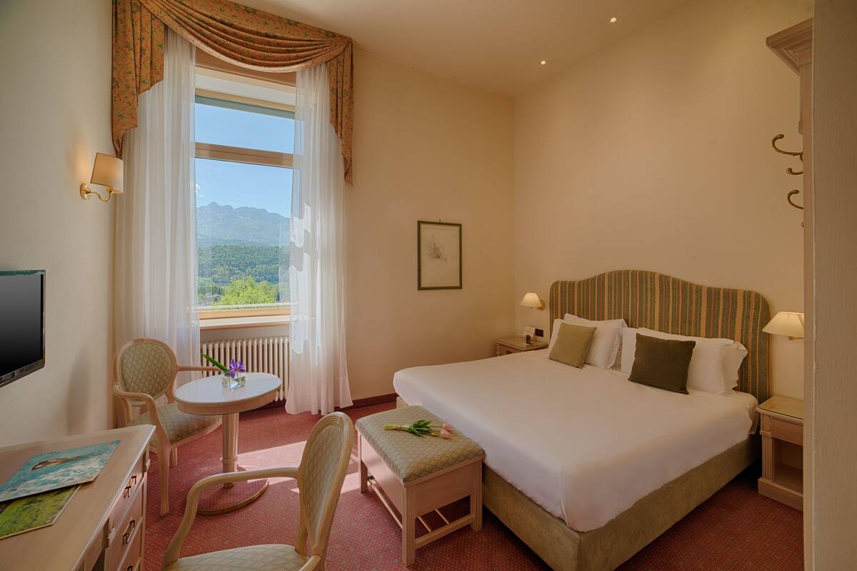 Ein Hotelzimmer mit einem großen Bett, einem Schreibtisch mit Stuhl, einem kleinen Tisch mit Vase und einem Fenster mit Vorhängen.