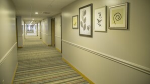 A hotel corridor with a striped carpet, framed botanical prints on the walls, and a long hallway leading to a door at the end. - Seffner Inn and Suites (Seffner)